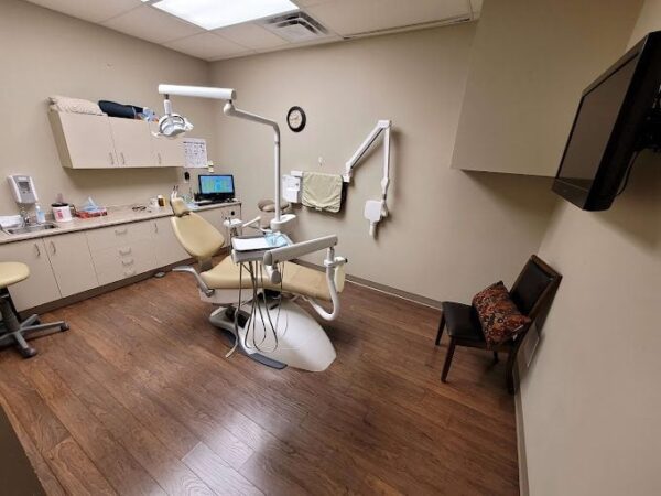 A dental office featuring a treatment chair, dental equipment, a monitor on a desk, and wooden flooring. A single chair is placed against the wall, and there are cabinets for storage. The room has warm lighting and a neutral color palette.