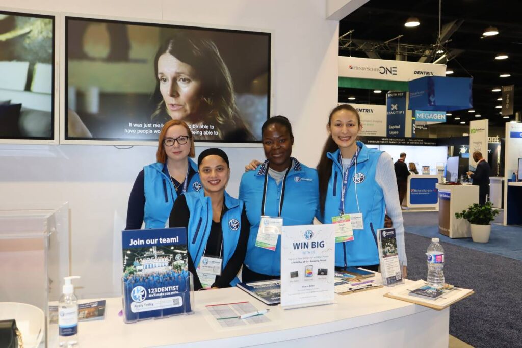 Four women in blue vests smile at the camera, standing behind a booth with promotional materials. A screen in the background displays a video featuring a woman. The setting appears to be a conference or trade show.