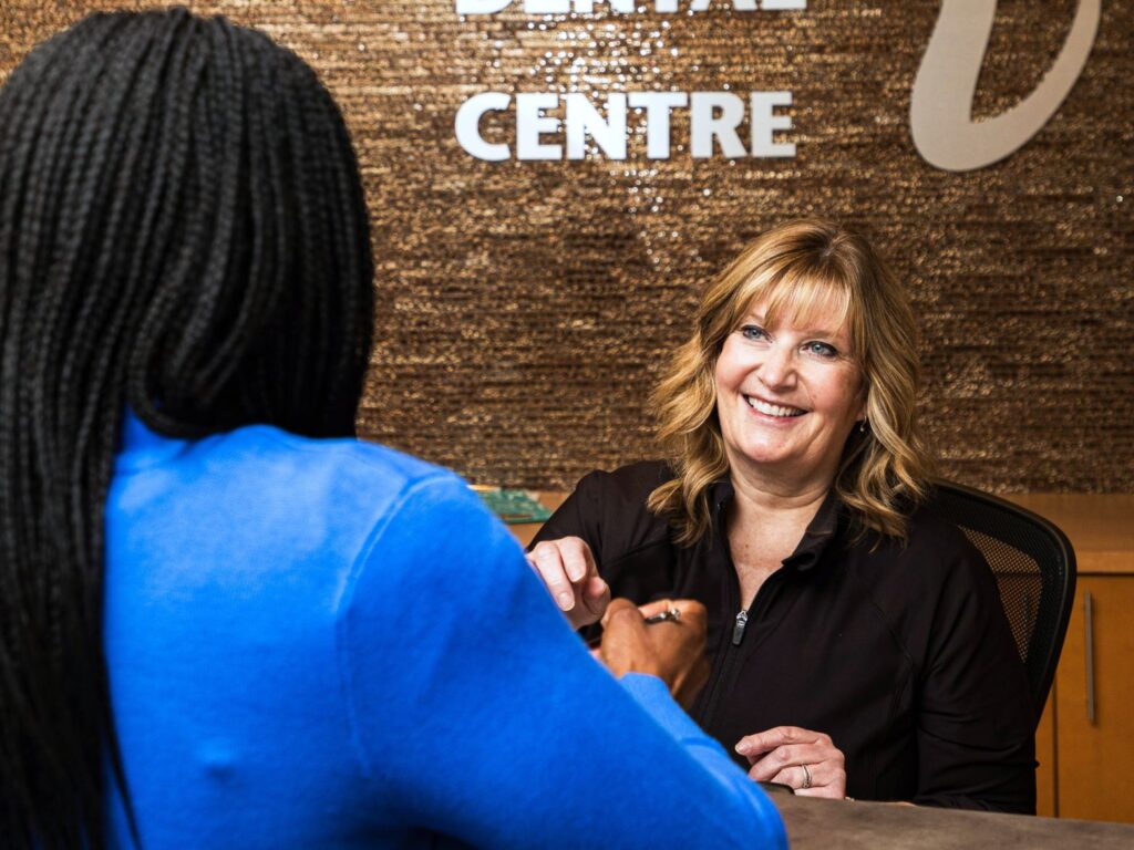 A friendly receptionist at a dental office smiles while interacting with a client. The setting is warm and welcoming, featuring a decorative wall with "Dental Centre" displayed.