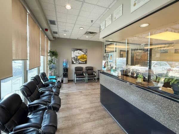 A modern dental office reception area featuring comfortable black chairs, large windows, and a glass partition displaying plants. Brightly lit with wall art, the space promotes a welcoming atmosphere for patients. The friendly 123Dentist Smiley adds a cheerful touch.