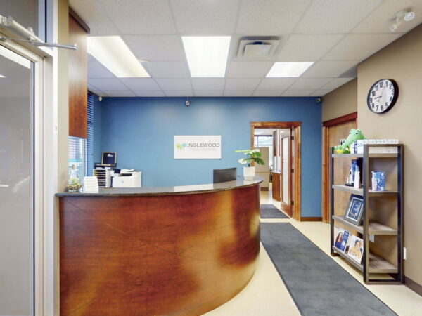 A modern reception area featuring a curved wooden desk, a blue accent wall, and a shelf with brochures. There is a clock on the wall and office equipment visible in the background, creating a welcoming and professional atmosphere.