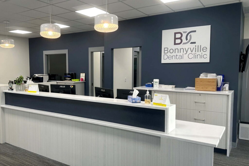 A modern dental clinic reception area featuring a white counter, blue walls, and a sign reading "Bonnyville Dental Clinic." The space is well-lit, with stylish light fixtures and organized workstations in the background.