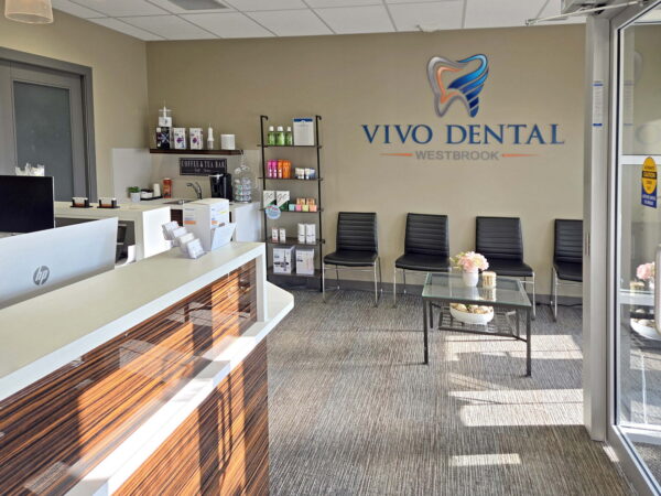 A dental clinic reception area featuring a modern desk, waiting chairs, and a display shelf with dental products. The wall prominently displays the "Vivo Dental" logo. Natural light illuminates the space, creating a welcoming atmosphere.