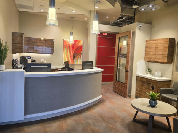 A modern reception area featuring a curved front desk, stylish decor, and warm lighting. The space includes a small seating area with a plant and artwork on the walls, creating an inviting atmosphere.