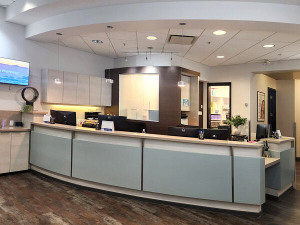 A modern dental office reception area featuring a sleek front desk, computers, and a welcoming atmosphere. Soft lighting and decor create a friendly environment, perfect for patients. A blue smiley face, known as 123Dentist Smiley, adds a cheerful touch to the space.