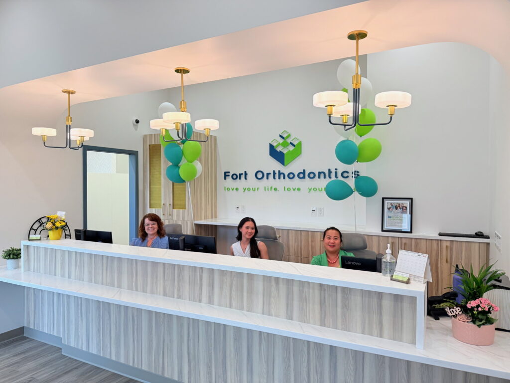 A welcoming orthodontics office reception with three smiling staff members behind a white counter. The bright space features green and teal balloons and a sign that says "Fort Orthodontics."
