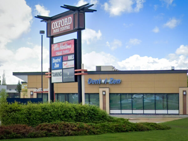 A modern dental office exterior is shown, featuring large windows and a prominent sign that reads "Oxford Park Centre." Nearby signage indicates various businesses, and greenery surrounds the building, contributing to a welcoming atmosphere. The image conveys a professional and approachable environment for dental care.