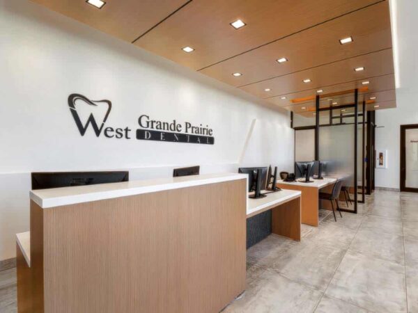 A modern dental office reception area featuring a sleek wooden desk, computers, and a wall sign that reads "West Grande Prairie Dental." The space is well-lit and has a contemporary design with neutral colors.