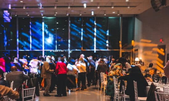 A lively event is taking place in a spacious hall with colorful lights illuminating the scene. Attendees gather and dance, enjoying the festivities, while tables are set for dining. In the background, large windows allow for a view of the vibrant atmosphere outside, creating a welcoming ambiance.