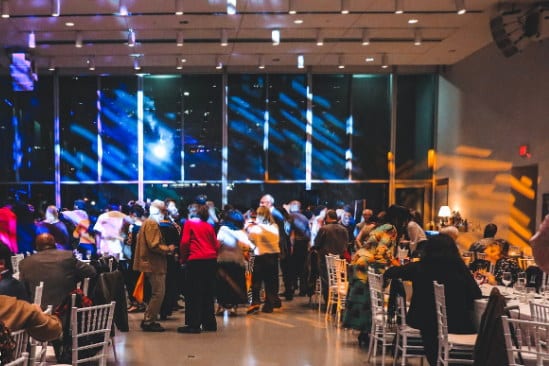 A lively event is taking place in a spacious hall with colorful lights illuminating the scene. Attendees gather and dance, enjoying the festivities, while tables are set for dining. In the background, large windows allow for a view of the vibrant atmosphere outside, creating a welcoming ambiance.