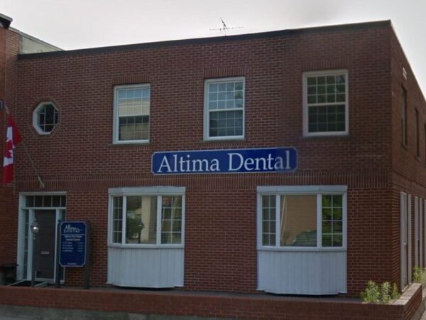 A brick building featuring the sign for Altima Dental. It has multiple windows, a doorway, and is situated next to another establishment. A Canadian flag is visible in front.