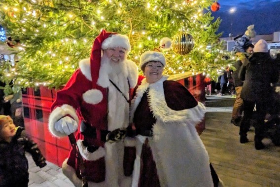 A cheerful Santa Claus and a woman dressed in a festive outfit pose together in front of a beautifully decorated Christmas tree, surrounded by holiday lights and a crowd of people enjoying the festive atmosphere.