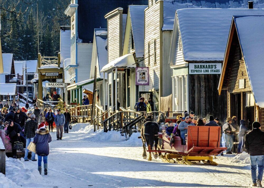 A bustling winter street scene with snow-covered buildings and people shopping, walking, and enjoying outdoor activities. Sleds are present, and the atmosphere captures a lively, festive spirit in a historic town.