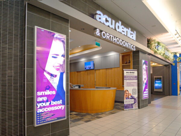 A dental and orthodontics clinic entrance featuring a modern design, with a reception area visible. Promotional banners display a smiling woman and the tagline "smiles are your best accessory." The setting is bright and welcoming, located within a shopping center.