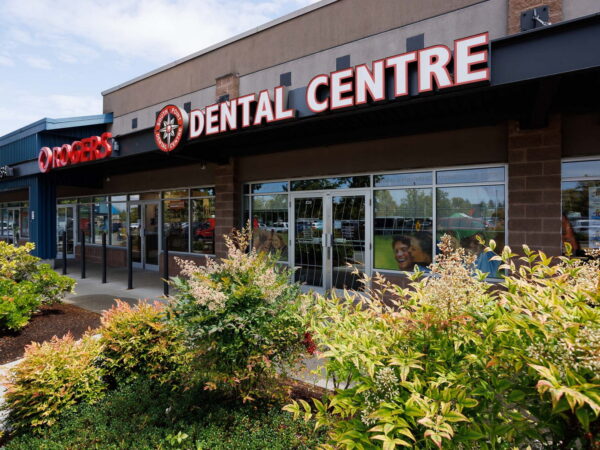A dental center building with a prominent sign that reads "DENTAL CENTRE," surrounded by shrubs and greenery. The entrance features large glass windows, providing a view into the office.