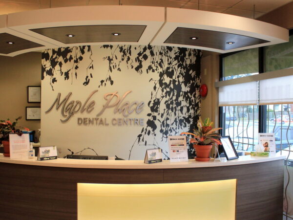 A modern dental office reception area featuring a stylish front desk and a decorative, nature-themed wall. The name "Maple Place Dental Centre" is prominently displayed, along with plants and informational materials for visitors. Natural light flows in through large windows.