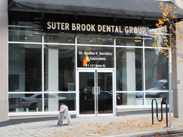 The image shows the entrance of Suter Brook Dental Group, featuring large glass doors and a sign displaying the name and doctor. The area has autumn leaves on the ground, indicating the season.