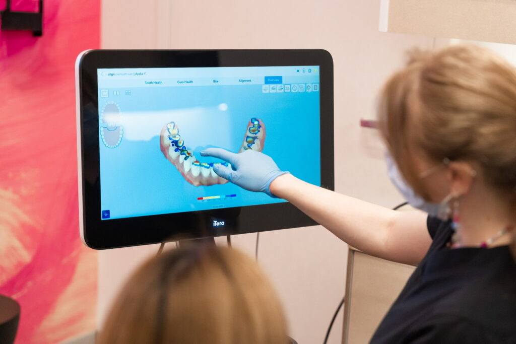 A dental professional, wearing gloves and a mask, is pointing at a digital screen displaying a dental model while consulting with a patient. The background has a colorful design, creating a friendly atmosphere in a dental office.