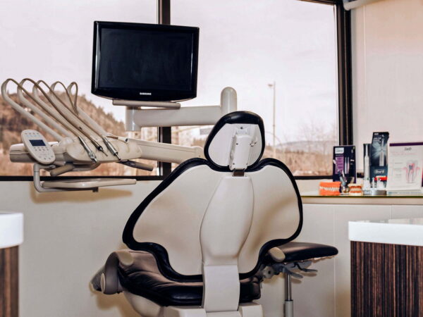 A dental examination room featuring a modern dental chair, overhead dental tools, and a flat-screen monitor. The large window provides natural light, enhancing the calming atmosphere of the space.