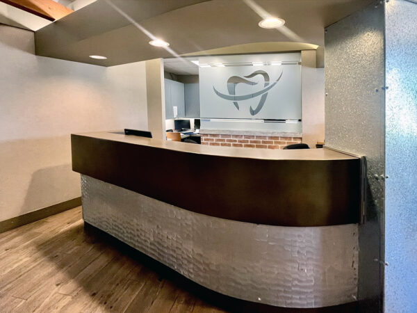 A modern dental reception area featuring a wooden front desk with a textured design. The background includes a logo on the wall and a warm, inviting atmosphere. The space is well-lit and organized, ready to welcome patients.