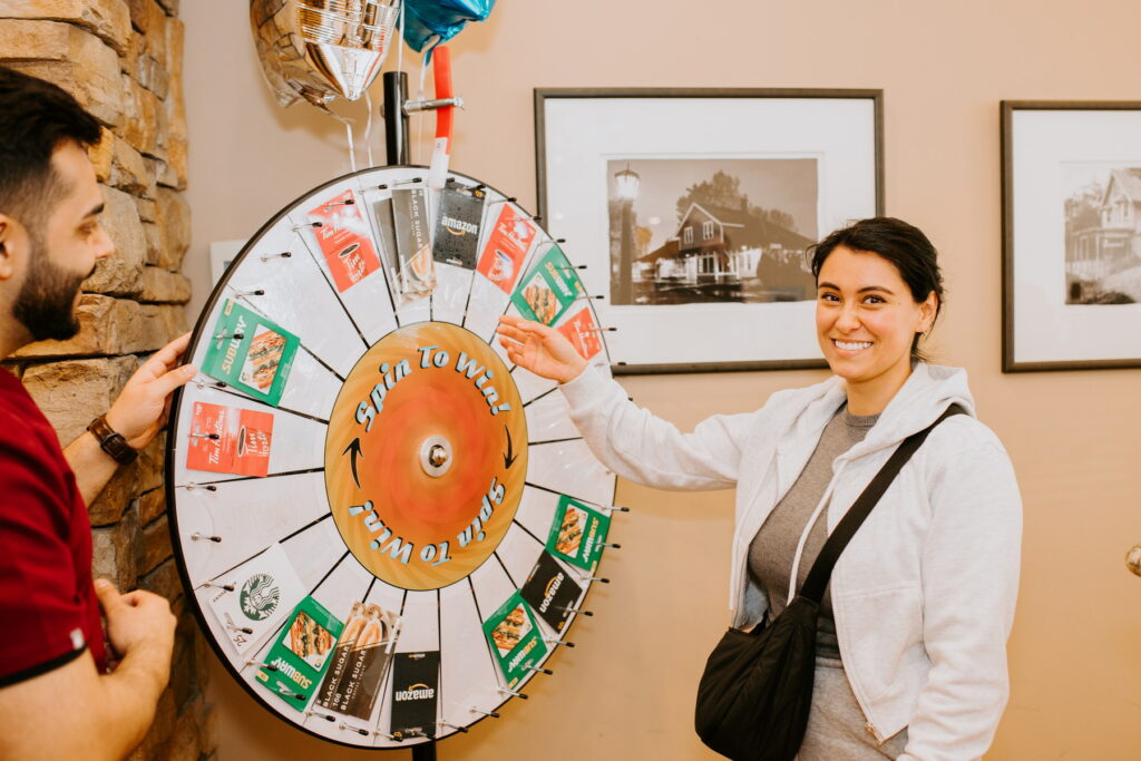 A woman smiles as she prepares to spin a colorful prize wheel while standing next to a man. The setting features framed photographs on the wall, creating a lively atmosphere. The 123Dentist Smiley can be seen in the background, adding a friendly touch to the scene.