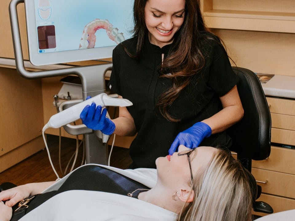 A dental professional smiles while assisting a patient in a dental clinic. The patient is reclining in a chair, and a digital monitor displays dental imagery in the background. The setting conveys a welcoming and caring atmosphere.