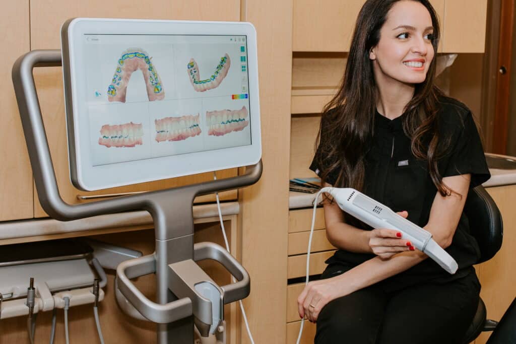 A dental professional sits in a clinic, smiling while holding a digital scanning tool. Behind her, a screen displays colorful dental images and charts, highlighting her focus on modern dental technology and care.