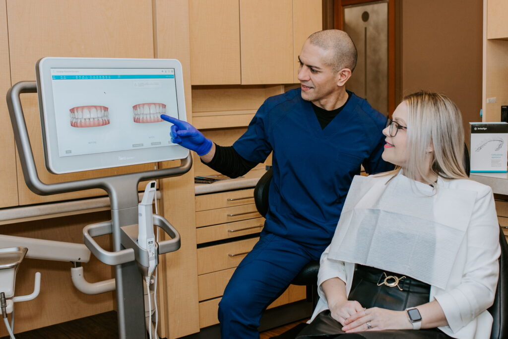 A dentist in scrubs points to a digital screen displaying dental images while discussing them with a patient seated in a dental chair. The office setting is modern and inviting, emphasizing patient care and communication. The 123Dentist Smiley adds a friendly touch to the environment.