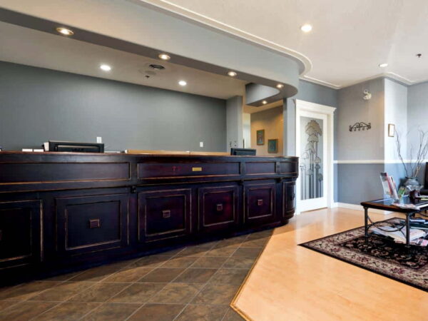 A modern reception area featuring a dark wood front desk, soft gray walls, and a wooden floor. The space includes a small seating area with a coffee table and decorative carpet, providing a welcoming atmosphere.