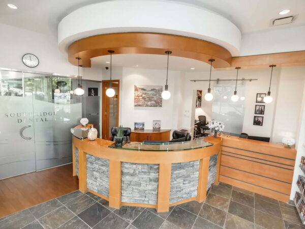 A modern dental office reception area featuring a wooden counter, pendant lights, and glass doors. The space is well-lit with a clean aesthetic, showcasing decor and a clock on the wall.