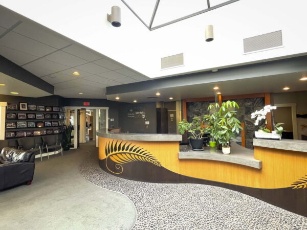 Interior view of a modern reception area featuring a curved front desk with plants, a cozy seating area, and decorative flooring. The space is well-lit with large windows and a skylight overhead. Walls are adorned with framed photos, creating a welcoming atmosphere.