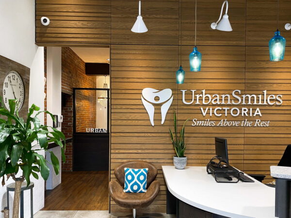 Reception area of Urban Smiles Victoria, featuring a wooden wall, stylish lighting, and a modern desk with a plant. A blue cushion adds a pop of color to the seating. The logo emphasizes a welcoming dental environment.