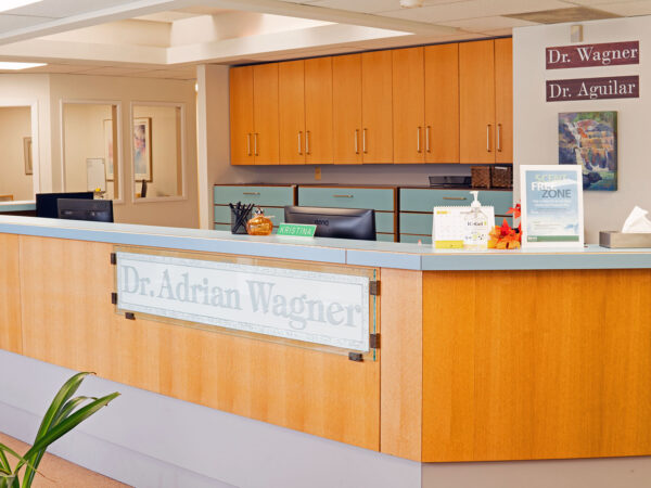 A modern dental reception area featuring a wooden front desk with the name "Dr. Adrian Wagner" prominently displayed. The space is bright and welcoming, with light-colored walls and various informational displays. A friendly 123Dentist Smiley adds a cheerful touch.