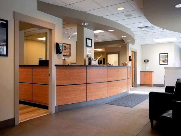 A modern reception area featuring a wooden front desk, neutral-colored walls, and a waiting area with black seating. The space is well-lit and organized, with decorative elements and functional areas for visitor check-in.
