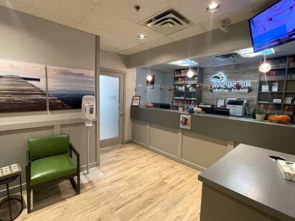 A modern dental office interior featuring a reception area with a green chair, a counter with dental supplies, and a scenic wall image. A television displays information, creating a welcoming atmosphere for patients. The friendly 123Dentist Smiley adds a cheerful touch.