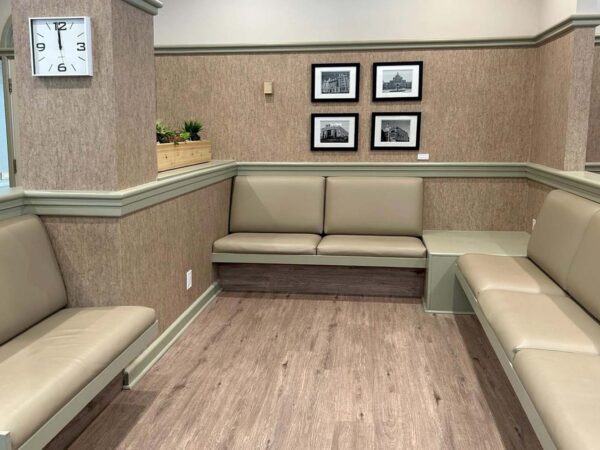 A modern waiting area featuring light brown benches along two walls, minimalistic decor, a wall clock, and black-and-white framed photos. The space has a clean, inviting atmosphere with wooden flooring and subtle green accents.