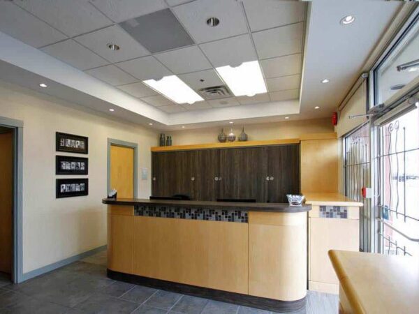 A modern reception area featuring a wooden front desk, muted lighting, and gray tile flooring. The walls are painted softly, with decorative elements and a large window providing natural light.
