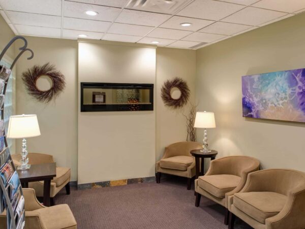A serene waiting area features two comfortable chairs and a small table, illuminated by two lamps. A decorative wall with artwork and two circular wall hangings complements a fish tank on a neutral-colored wall, creating a calm atmosphere. A brochure stand is positioned on the left.