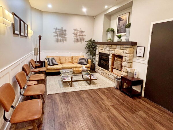 A cozy waiting area featuring a brown leather sofa, a coffee table, and several wooden chairs along the wall. A stone fireplace adds warmth, while modern artwork and indoor plants enhance the inviting atmosphere.
