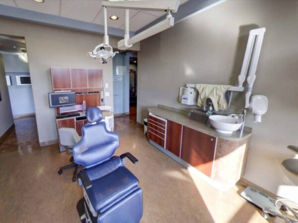 A modern dental office featuring a blue dental chair, a sink, and various dental equipment. The interior is well-lit with wooden cabinets and a clean, organized layout, creating a professional atmosphere for patient care.