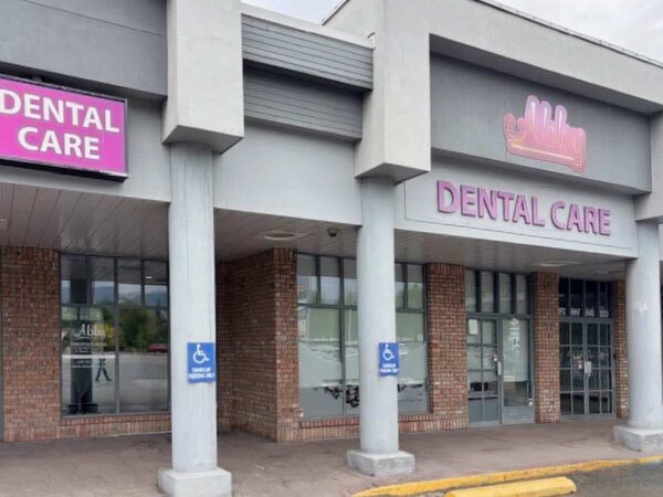 Exterior of a dental clinic featuring the sign "DENTAL CARE" in large pink letters. The building has large windows and accessible parking signs in front. The structure has a modern look with a mix of brick and concrete elements.