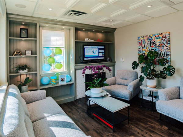 A modern waiting area featuring light-colored sofas, a TV, and vibrant artwork. There are shelves with decor and plants, creating a welcoming atmosphere. Bright windows illuminate the space, complemented by a friendly 123Dentist Smiley.