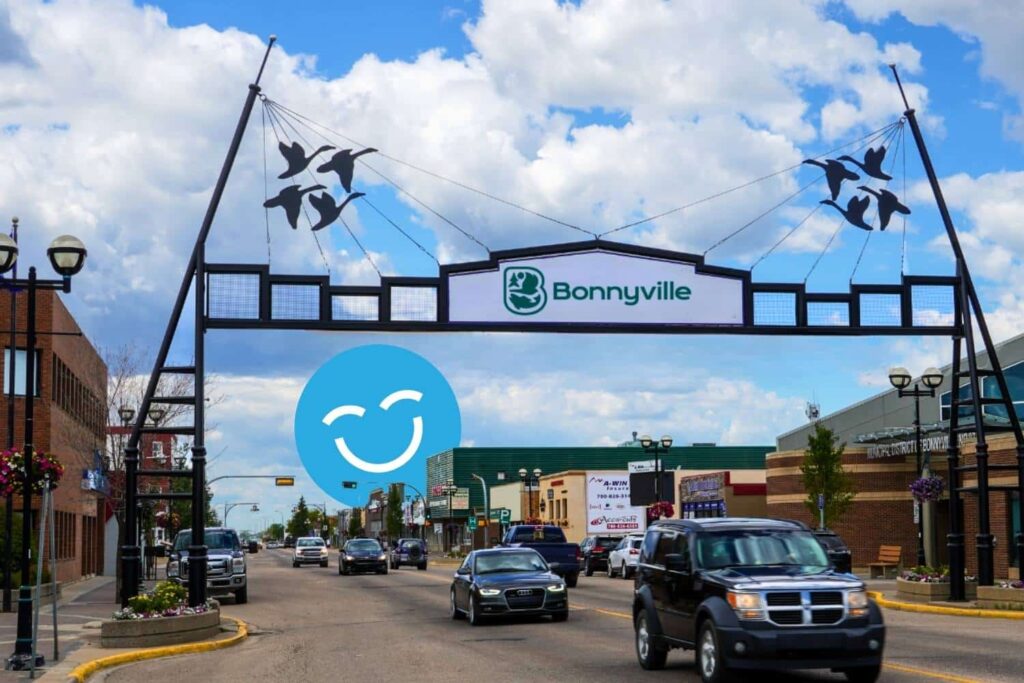 A street scene in Bonnyville, featuring a welcoming archway with the town's name and artistic representations of birds above. Vehicles are driving along a busy road under a partly cloudy sky, with shops visible on either side.