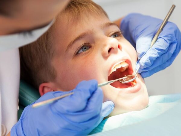 A dentist examines a young boy's teeth, using dental tools. The boy is seated in a dental chair, with a light shining on his open mouth. The dentist wears blue gloves and a mask.