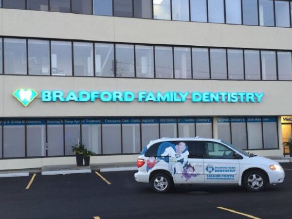 A dental clinic building with a sign reading "Bradford Family Dentistry" in bright blue letters. A vehicle with a dental logo is parked nearby, along with several empty parking spaces. The setting appears to be a professional and welcoming environment.
