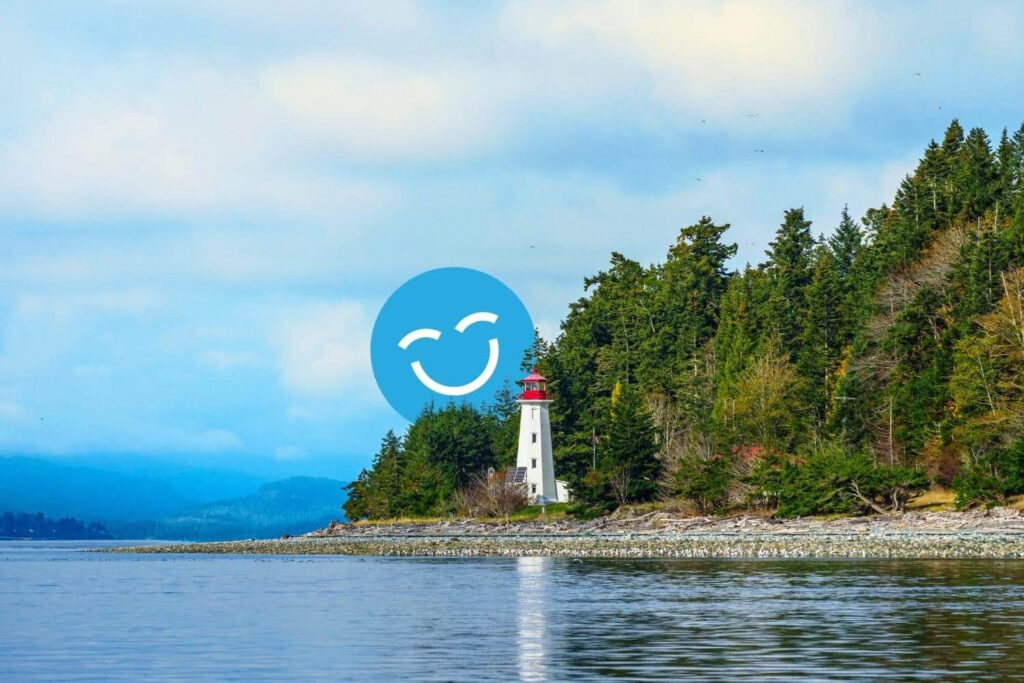 A lighthouse stands on a rocky shore surrounded by dense green trees, with calm water in the foreground and a partly cloudy blue sky. A blue, smiling emoji with closed eyes is superimposed in the sky.