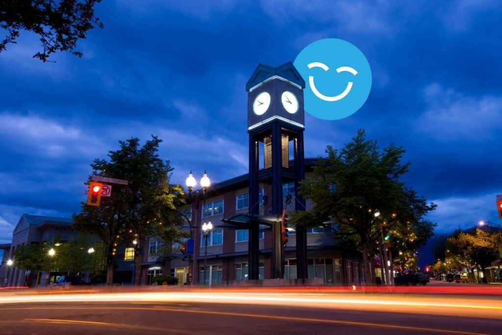 Clock tower illuminated at dusk with a smiling face in the sky, surrounded by trees and a street with light trails.