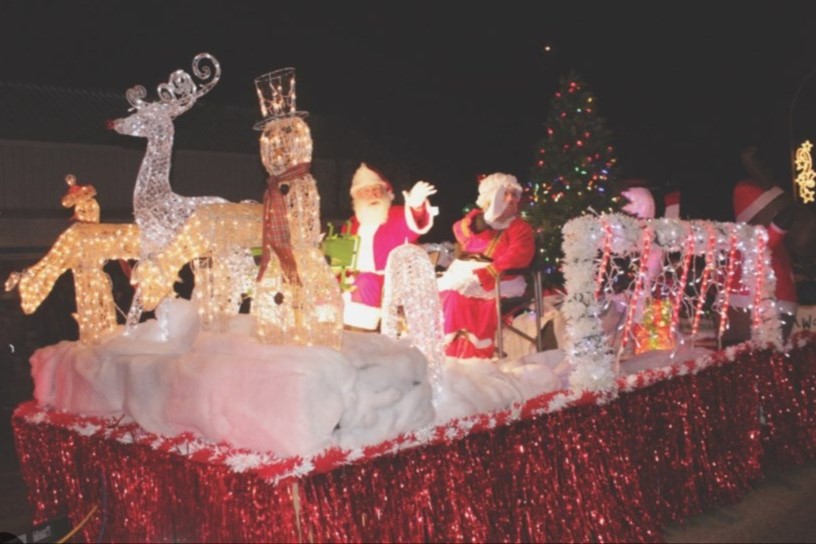 A festive parade float decorated with Christmas lights features Santa Claus, a reindeer, a nutcracker, and a snowy backdrop. The float is adorned with red glittery material and a Christmas tree, creating a cheerful holiday scene at night.