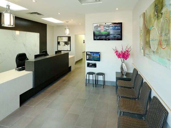 A modern reception area featuring a sleek front desk, comfortable seating, and decorative artwork. There is a television mounted on the wall displaying images, with flowers adding a touch of color to the inviting space.