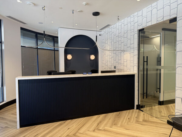A modern dental reception area featuring a sleek black counter, stylish lighting, and large windows. The design showcases a mix of contemporary elements with a warm, inviting atmosphere. A friendly blue smiley face, the 123Dentist Smiley, adds a cheerful touch to the space.